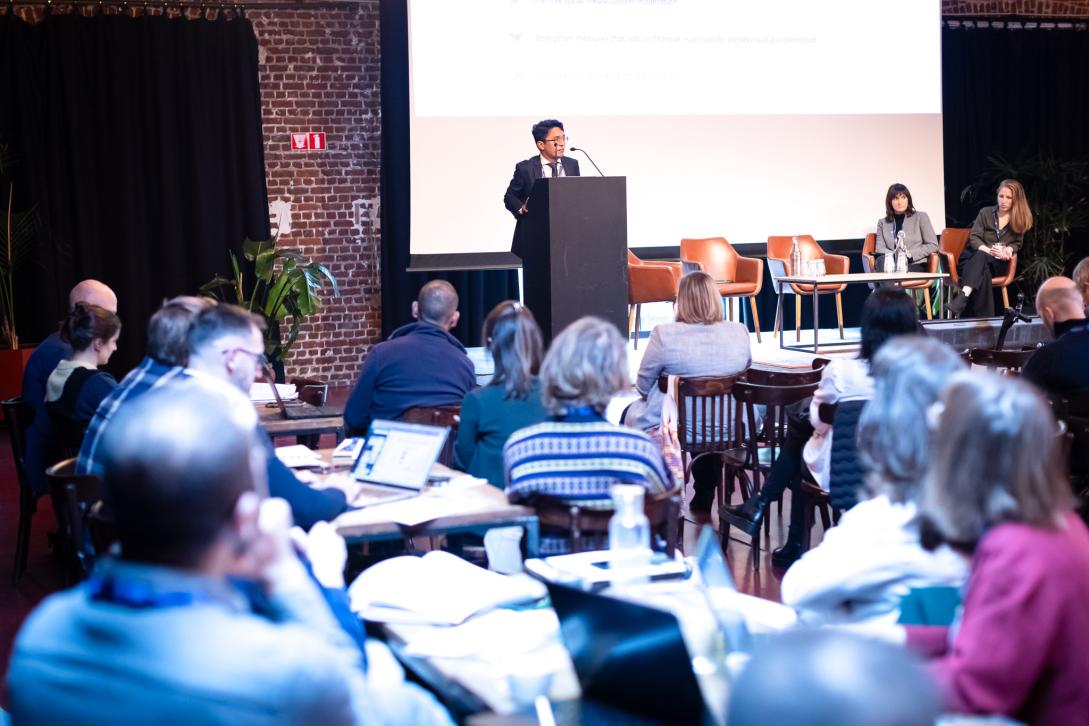 Picture from the event: speaker in the podium, two other speakers sitting beside him and the audience